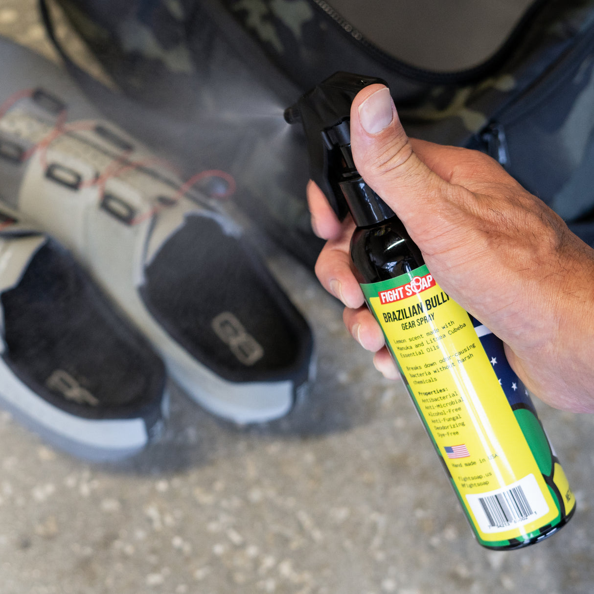 A hand holding a spray bottle labeled "Brazilian Bully Gear Spray" with a black nozzle, near a pair of sneakers.