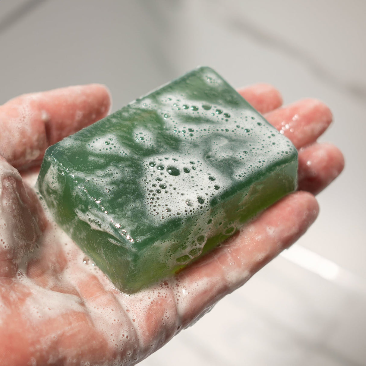 A hand holding a rectangular, green soap bar with bubbles and foam on its surface, labeled "TRIPLE THREAT.