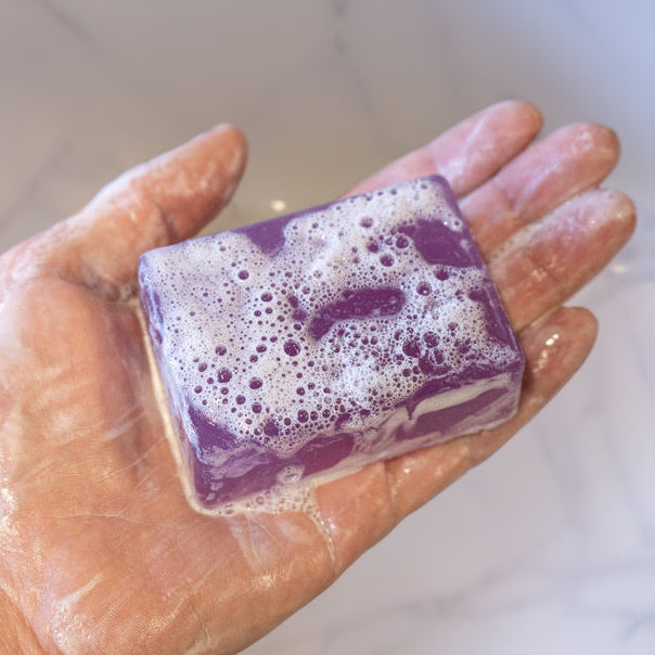 A hand holding a purple soap bar labeled VICIOUS VIXEN, with soap suds and foam on the surface and fingers.