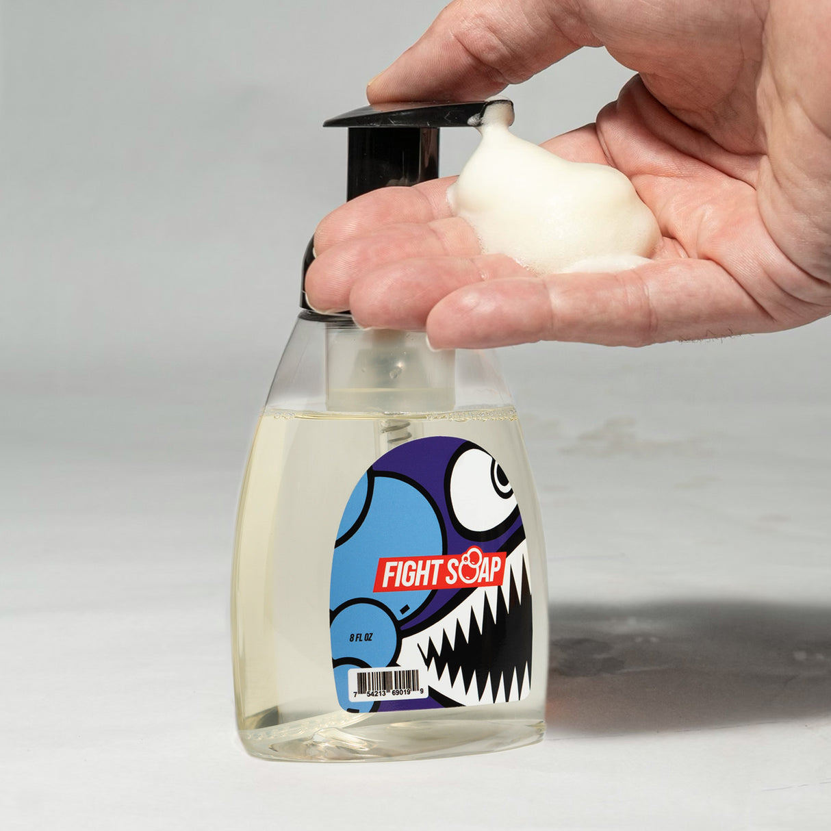 A hand dispenses foaming hand soap from a clear bottle with a cartoon monster label reading "Fight Soap.