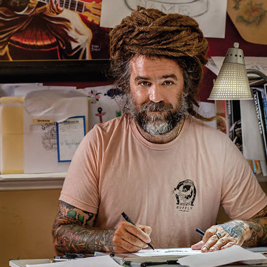 A tattoo artist with dreadlocks and tattoos on his arms, sitting at a desk with tattoo supplies, holding a marker.