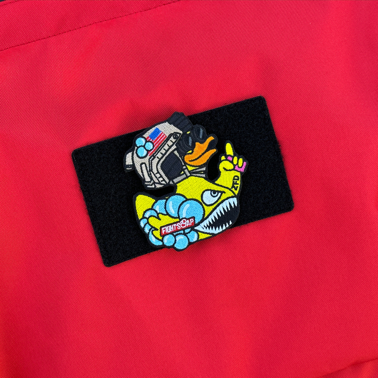 Colorful embroidered patch featuring a cartoon duck wearing a helmet, goggles, and holding a finger up, on a red fabric background.