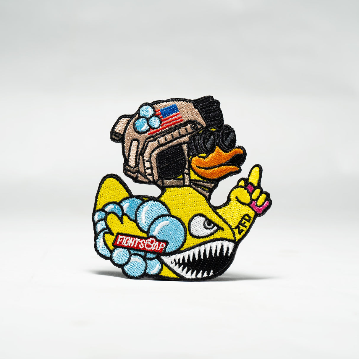 Colorful embroidered patch of a duck wearing goggles and a helmet, holding a shark with "Fight Soap" written on it, and giving a peace sign.