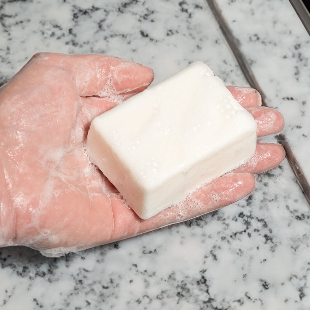 A hand holding a white soap bar labeled "LITTLE FRIEND" over a marble countertop.